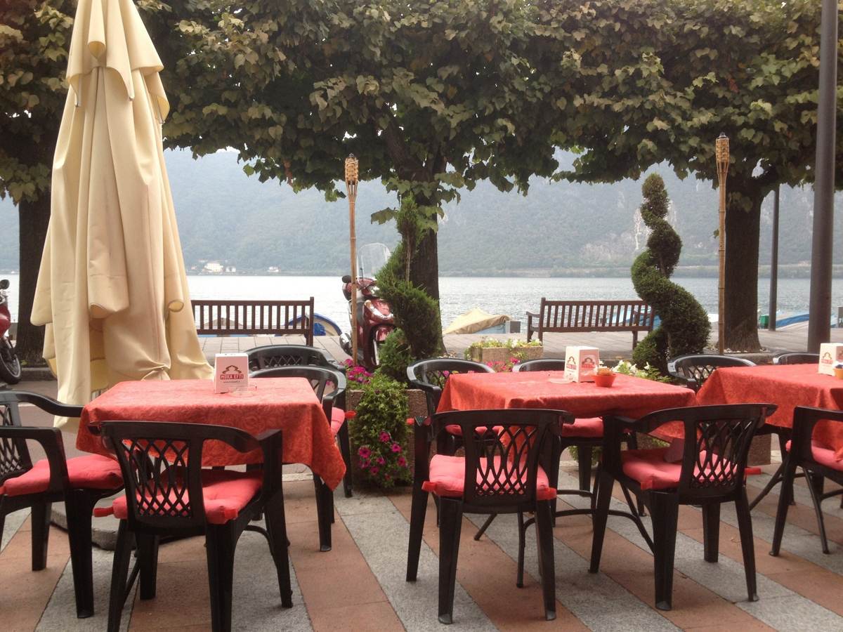 A lakeside restaurant terrace with red tables and umbrellas at Lake Lugano