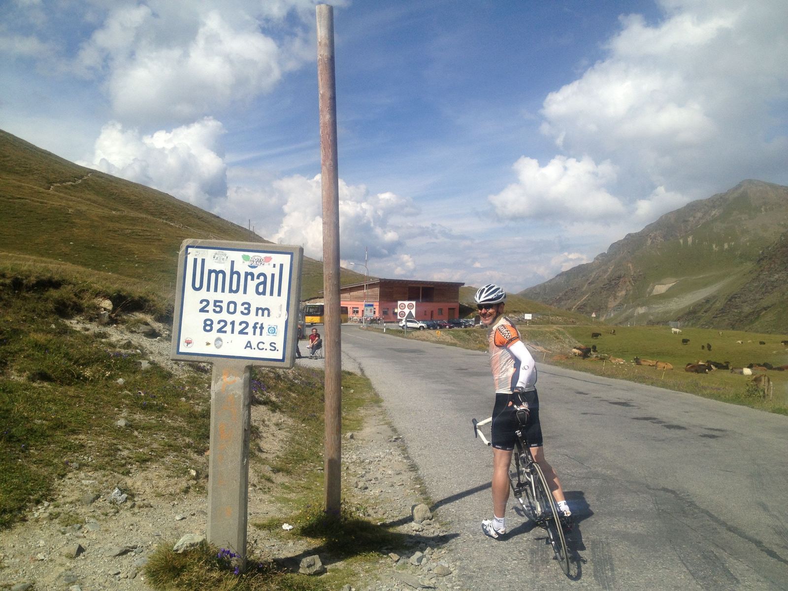 Umbrail Pass 2503m altitude sign with Kevin and his bike, cows grazing in the background
