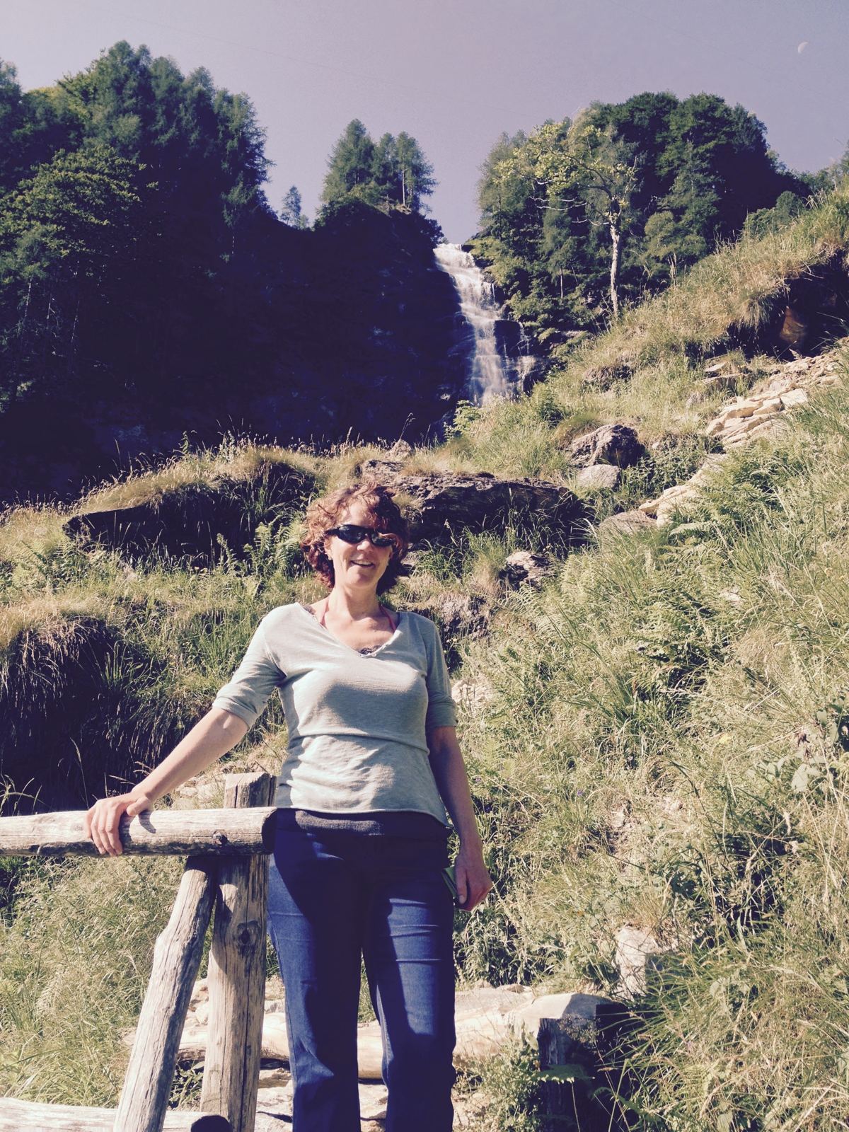 Kevin's wife standing by a wooden fence on a mountain trail near a waterfall above Sonogno
