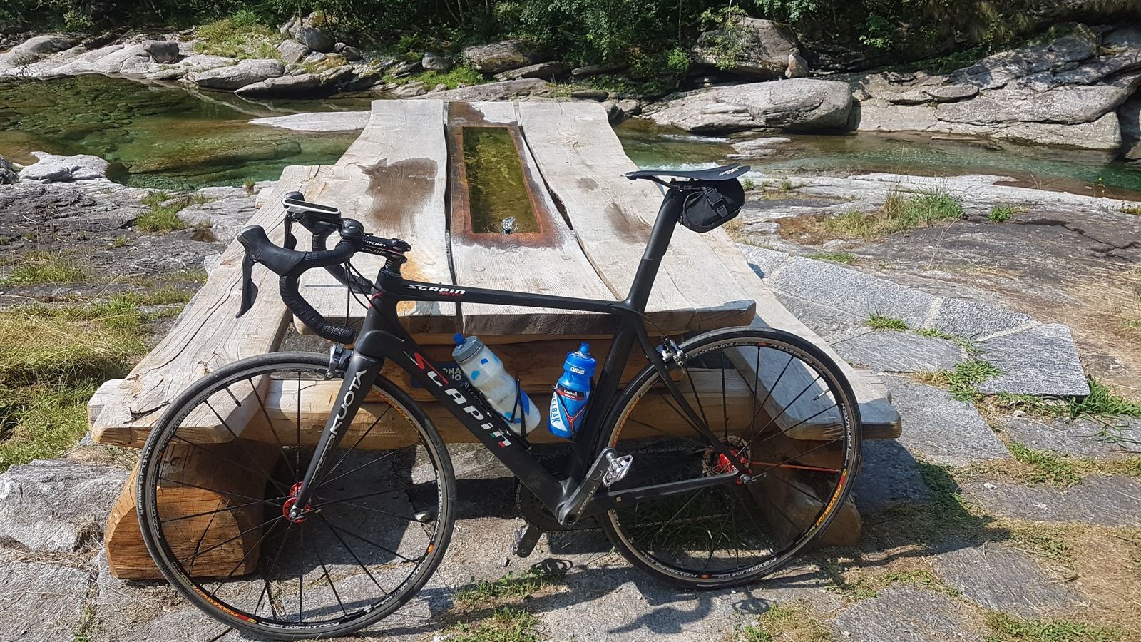 A road bike resting on a wooden table beside an emerald swimming pool in the Verzasca river